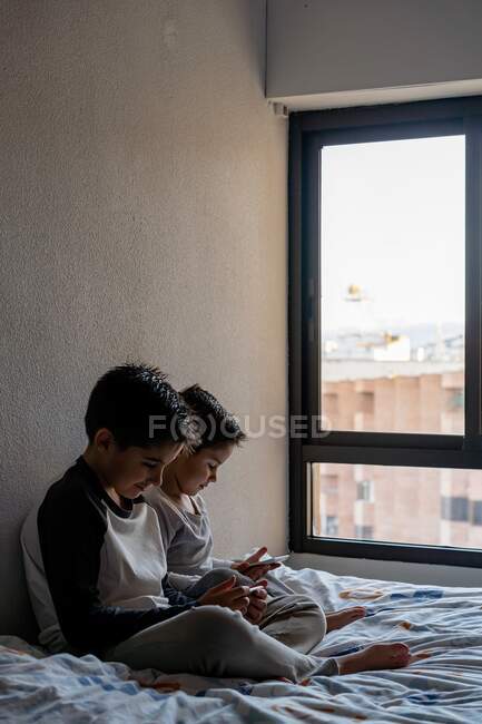 Hermanos sentados en una cama acogedora y viendo dibujos animados en los teléfonos celulares mientras se relajan en casa - foto de stock
