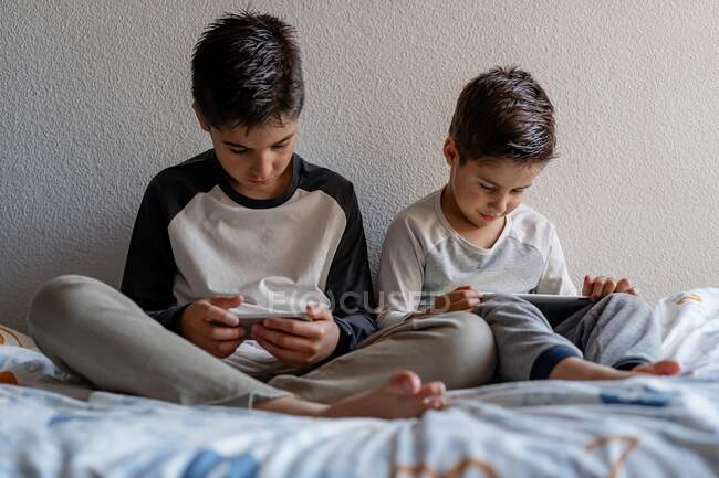 Brothers sitting on cozy bed and watching cartoons on cellphones while relaxing at home — Stock Photo