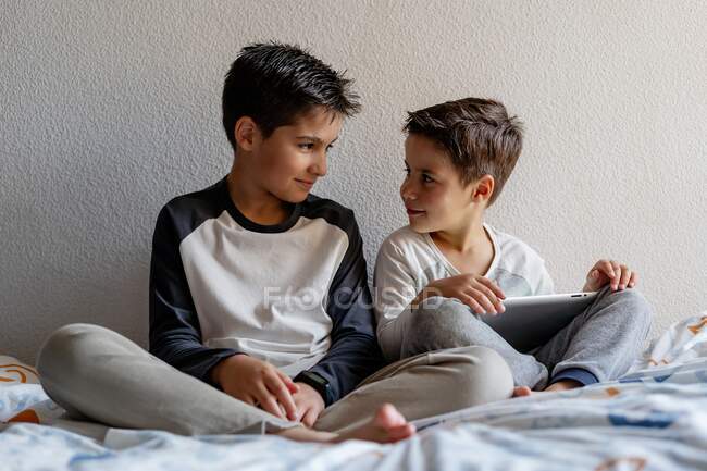 Smiling brothers in sleepwear sitting on cozy bed and watching movie together while entertaining at home — Stock Photo