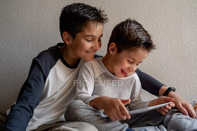 Lächelnde Brüder in Schlafanzügen sitzen auf einem gemütlichen Bett und schauen gemeinsam Filme, während sie zu Hause unterhalten werden — Stockfoto