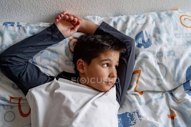 Von oben ein gelassenes Kind in Freizeitkleidung, das auf einem bequemen Bett liegt und wegschaut, während es am Wochenende zu Hause chillt — Stockfoto