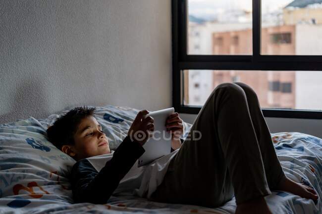 Niño encantado usando tableta en el dormitorio durante el fin de semana - foto de stock