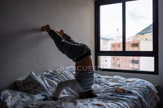 Rückansicht des niedlichen Kindes im Pyjama, das am Wochenende Spaß hat und Kopfstand auf weichem Bett zeigt, während es sich an die Wand lehnt — Stockfoto
