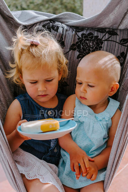 High angle of cute sisters sitting in hammock in courtyard with plate of homemade ice lolly — Stock Photo