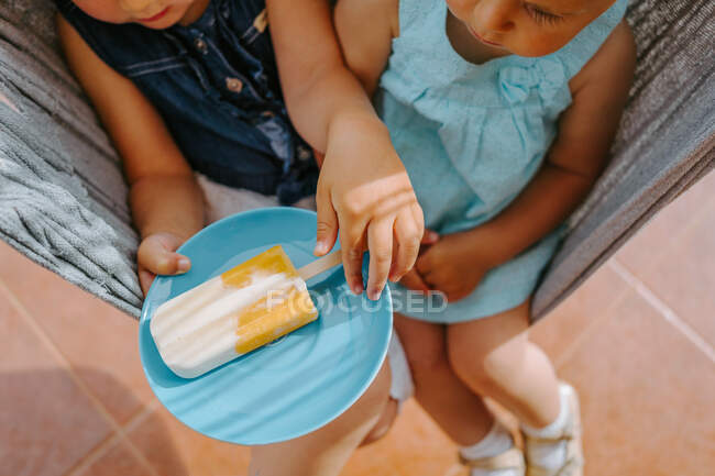 High angle of cropped unrecognizable cute sisters sitting in hammock in courtyard with plate of homemade ice lolly — Stock Photo