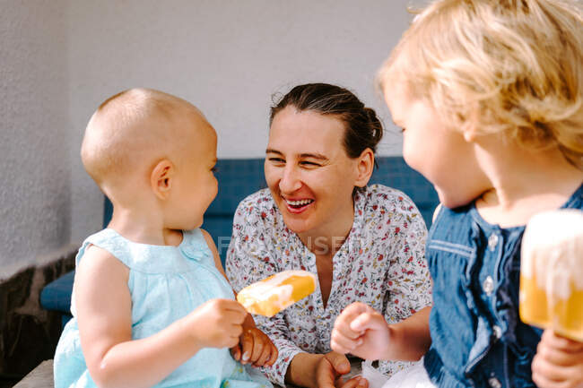 Веселая женщина и маленькие сестры сидят во дворе и наслаждаются домашним мороженым, глядя друг на друга — стоковое фото