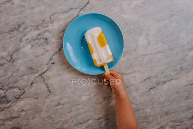 Vista superior de la mano de niño irreconocible recortado agarrando una deliciosa paleta de fruta colocada en la mesa - foto de stock