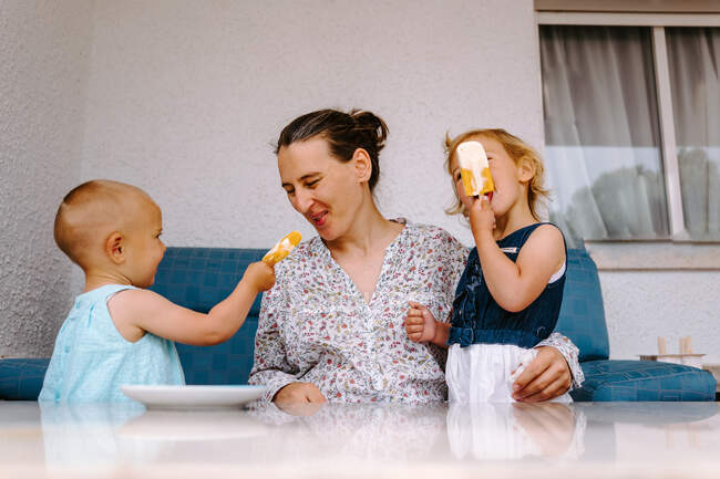 Fröhliche Frau und kleine Schwestern sitzen im Hof und genießen selbstgemachtes Eis am Stiel, während sie einander anschauen — Stockfoto