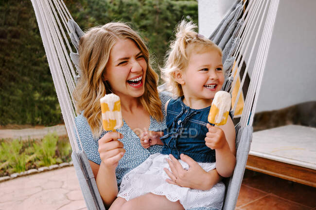Mãe e filha alegres abraçando na rede no terraço com saborosos piruetas de gelo e desfrutando de verão juntos — Fotografia de Stock