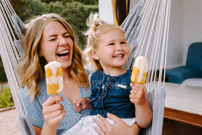 Mère et fille joyeux câlins dans un hamac sur la terrasse avec des sucettes de glace savoureuses et profiter de l'été ensemble — Photo de stock