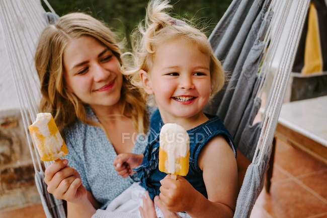 Cheerful mother and daughter hugging in hammock on terrace with tasty ice lollies and enjoying summer together — Stock Photo