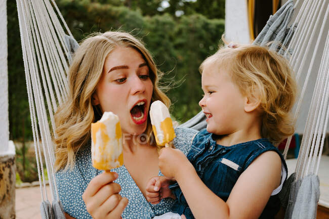 Mãe e filha alegres abraçando na rede no terraço com saborosos piruetas de gelo e desfrutando de verão juntos — Fotografia de Stock
