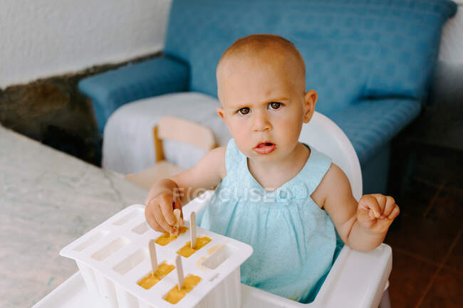 Ângulo alto de bebê bonito sentado em cadeira alta no terraço e comer deliciosos picolés enquanto olha para a câmera — Fotografia de Stock