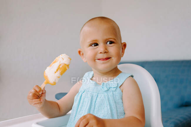 Lindo niño sentado en la silla alta en la terraza y comiendo deliciosos paletas mientras mira hacia otro lado - foto de stock