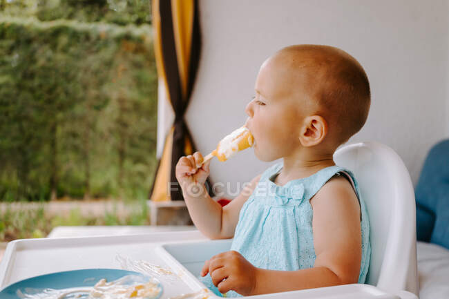 Nettes Kleinkind sitzt auf dem Hochstuhl auf der Terrasse und isst köstliche Eis am Stiel, während es wegschaut — Stockfoto