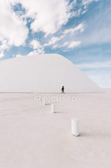 Невпізнавана людина, що ходить по порожній площі біля білого вигнутого будинку Оскара Німеєра Міжнародного культурного центру в Астурії (Іспанія) в сонячні дні з синім хмарним небом на задньому плані. — стокове фото
