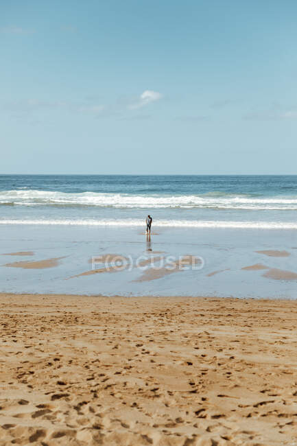 Vue arrière du voyageur anonyme debout sur la côte pittoresque de la mer près du sable avec des empreintes de pas sous le ciel nuageux par une journée ensoleillée — Photo de stock