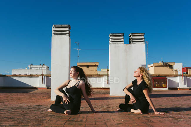Vista lateral da mulher de meia-idade com a filha jovem sentada em Ardha Matsyendrasana Variação pose enquanto praticam ioga juntos no terraço no terraço do telhado em dia ensolarado na cidade — Fotografia de Stock