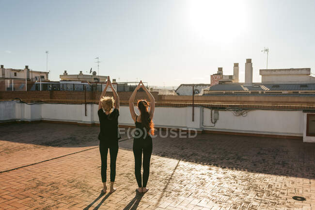 Полный назад вид зрелой женщины с молодой дочерью в черной спортивной одежде, стоящей в положении восходящего салюта с поднятыми руками во время практики йоги на крыше — стоковое фото