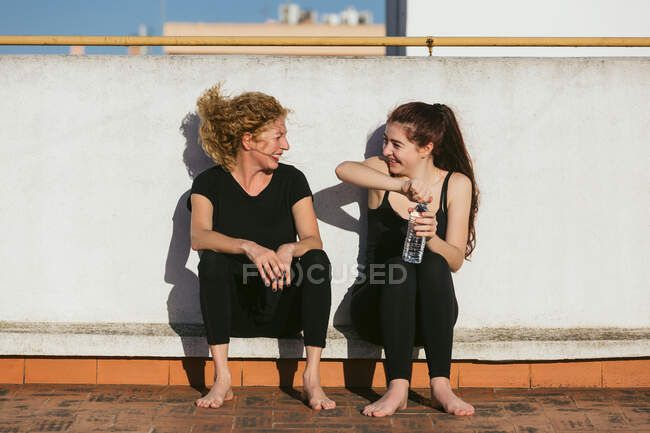 Joven alegre con madre de mediana edad en ropa deportiva hablando y riendo mientras se sienta con botella de agua en la azotea y descansando después de practicar yoga juntos - foto de stock