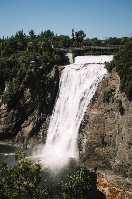 Pintoresco paisaje con solitaria puente mirador sobre cascada en el río que cae del acantilado rocoso entre el bosque denso verde bajo el cielo azul claro - foto de stock