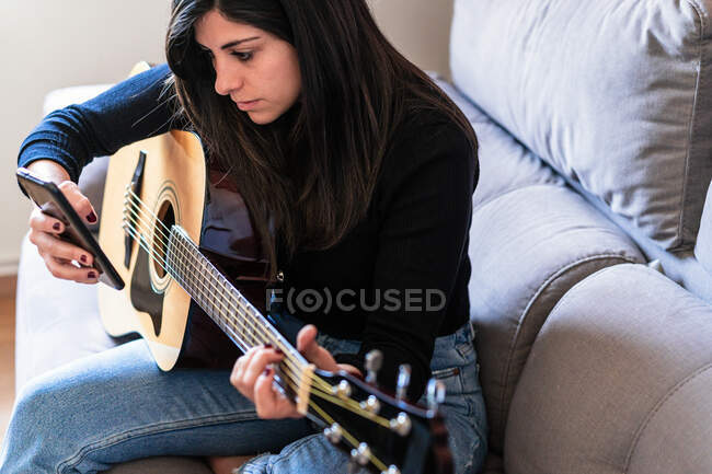 Mujer tocando la guitarra sentada en su sofá en casa y aprendiendo con lecciones en línea y algunas máscaras están colgando debido a la contención. Detrás de ella hay una pared de ladrillo - foto de stock