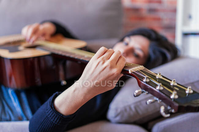 Женщина, играющая на гитаре, сидит дома на диване и учится с помощью онлайн-уроков и некоторых масок висят из-за сдерживания. За ним кирпичная стена. — стоковое фото