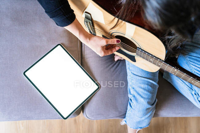 Donna che suona la chitarra seduta sul divano a casa e impara con lezioni online con un tablet digitale con uno schermo vuoto dall'alto — Foto stock