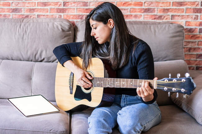 Женщина, играющая на гитаре, сидит дома на диване и учится с помощью онлайн-уроков с цифровым планшетом с цифровым планшетом с чистым экраном сверху — стоковое фото