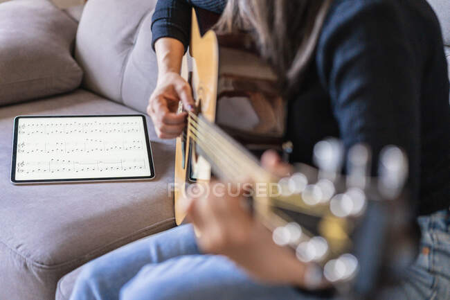 Donna che suona la chitarra seduta sul divano a casa e impara con lezioni online con un tablet digitale con uno schermo vuoto dall'alto — Foto stock