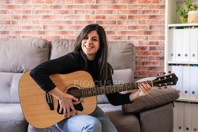 Femme jouant de la guitare assise sur son canapé à la maison et apprenant avec des leçons en ligne et certains masques sont suspendus en raison de confinement. Derrière il y a un mur de briques — Photo de stock