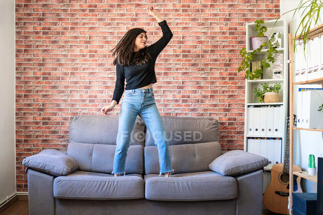 Молода жінка танцює на дивані у своєму будинку. За нею цегляна стіна — стокове фото