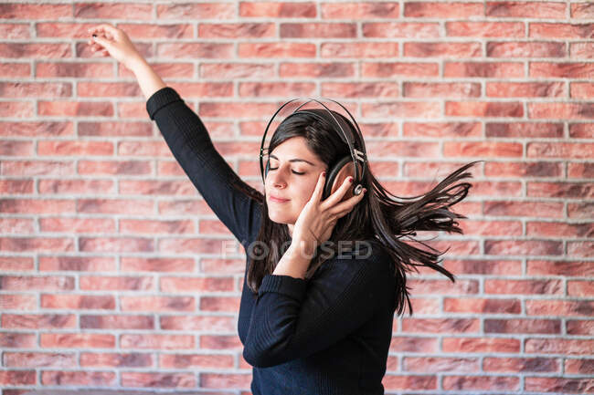 Mujer escuchando música en casa con auriculares vintage de madera. Detrás de ella hay una pared de ladrillo - foto de stock