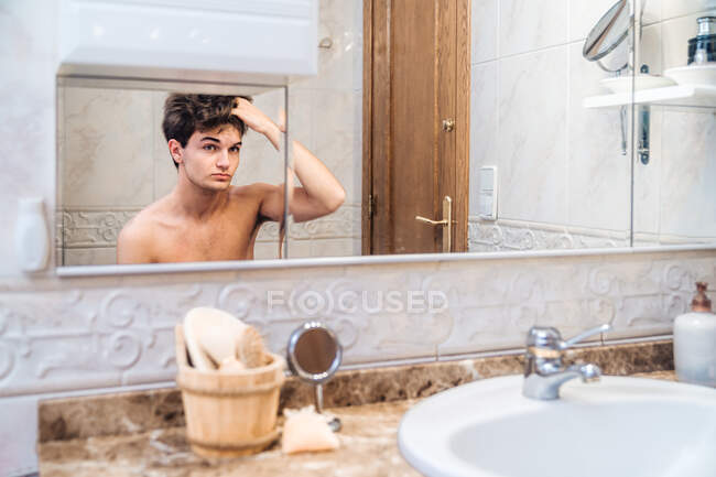 Красивый мужчина с обнаженным туловищем, стоящим в современной ванной комнате и смотрящим в зеркало утром — стоковое фото