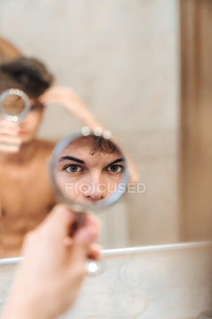 Varón serio de pie en el baño luminoso y mirando en espejo redondo por la mañana - foto de stock
