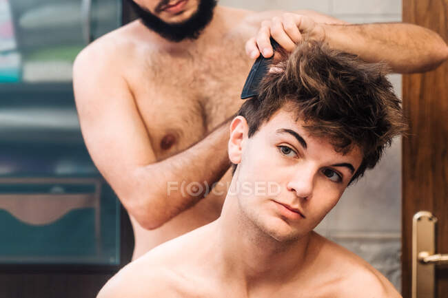 Homem de colheita escovando o cabelo do jovem e fazendo penteado no banheiro brilhante — Fotografia de Stock