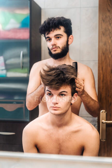 Homme brossant les cheveux du jeune homme et faisant coiffure dans la salle de bain lumineuse — Photo de stock
