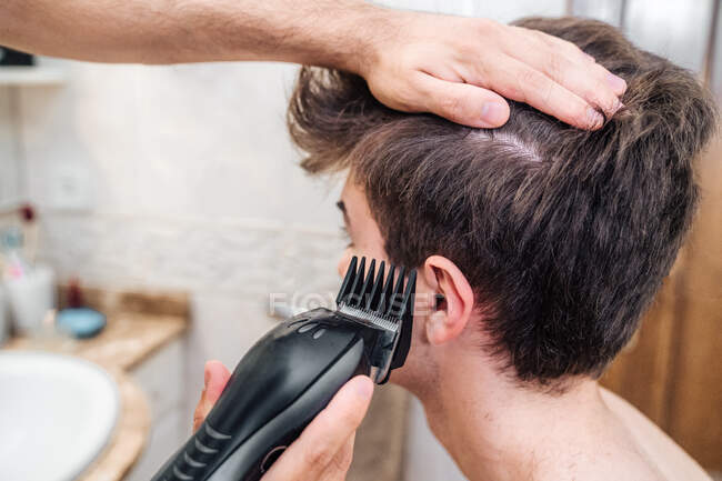 Мужчина с триммером для волос стрижет парня в современной ванной комнате дома — стоковое фото