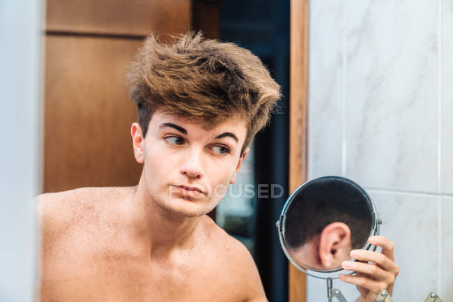 Calma ragazzo con torso nudo guardando se stessi in ulteriore piccolo specchio dopo auto tagliare i capelli in bagno leggero a casa — Foto stock