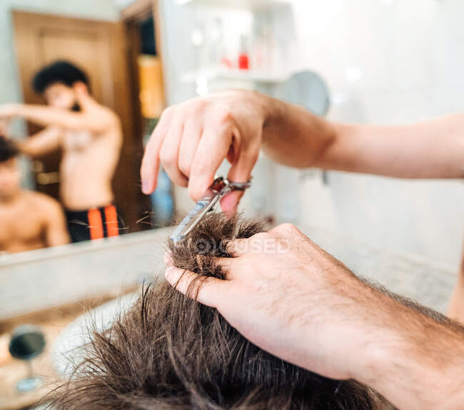 Вид ззаду невизначеного чоловіка, що робить зачіску хлопцеві, використовуючи ножиці проти розмитого інтер'єру легкої ванної кімнати вдома — стокове фото