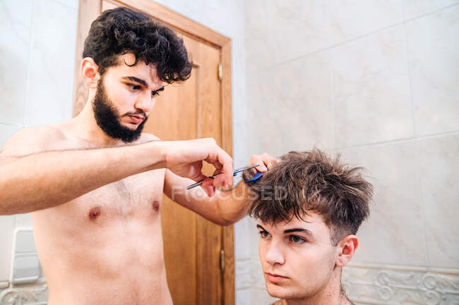 Male making haircut to guy using scissors against blurred interior of light bathroom at home — Stock Photo