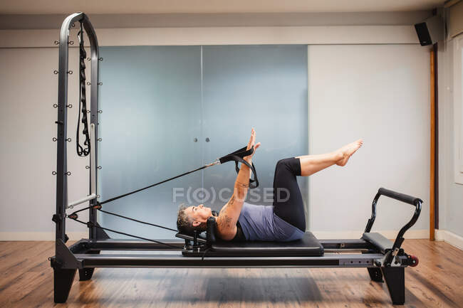 Vista lateral da fêmea adulta em sportswear deitado na máquina pilates e fazendo exercícios com bandas de resistência — Fotografia de Stock