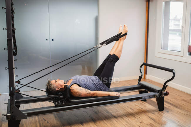Adult female in sportswear lying on pilates machine and stretching legs with resistance bands during training in modern gym — Stock Photo