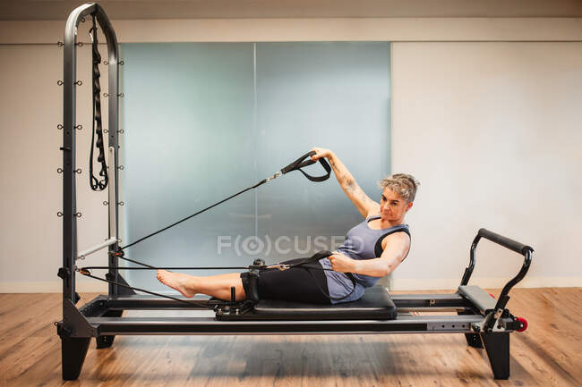 Seitenansicht einer erwachsenen Frau in Sportbekleidung, die auf einem Pilates-Gerät liegt und Übungen mit Widerstandsbändern macht — Stockfoto