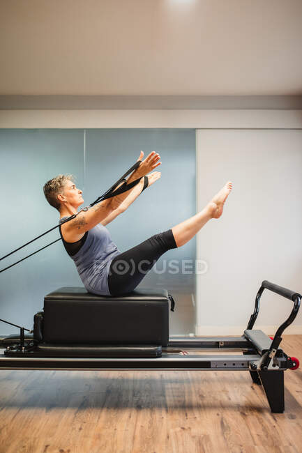 Vista laterale della femmina adulta in abbigliamento sportivo sdraiata sulla macchina pilates e facendo esercizi con bande di resistenza — Foto stock