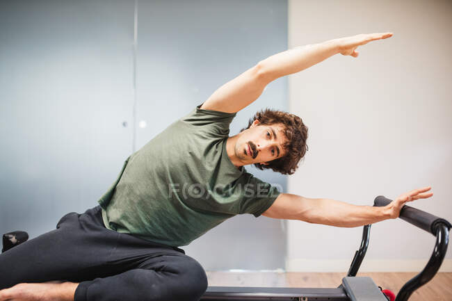 Atleta masculino focado em activewear sentado na máquina pilates e fazendo curvas laterais enquanto olha para a câmera durante o treinamento — Fotografia de Stock