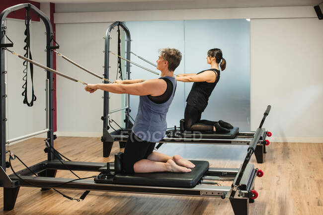 Side view of focused sportswomen in activewear doing exercises on pilates machine and pumping muscles with metal resistance equipment — Stock Photo
