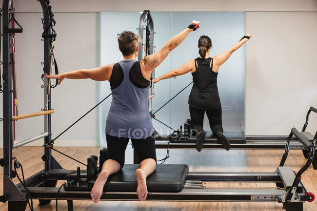 Vista posterior de las deportistas enfocadas en ropa deportiva haciendo ejercicios en la máquina de pilates y bombeando músculos con equipos de resistencia metálica - foto de stock