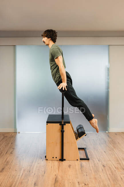 Вид сбоку сильного мужчины, балансирующего на руках на стуле пилатеса во время тренировки в современном спортзале — стоковое фото