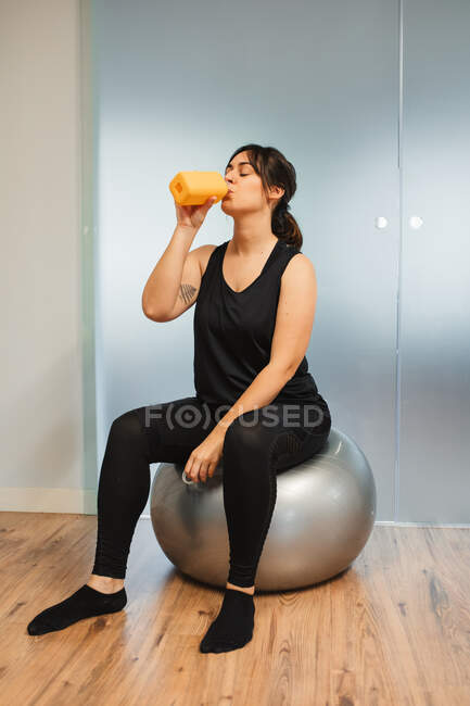 Mujer cansada en ropa deportiva sentada en la pelota de fitness y disfrutando del agua dulce después de un intenso entrenamiento en el gimnasio - foto de stock